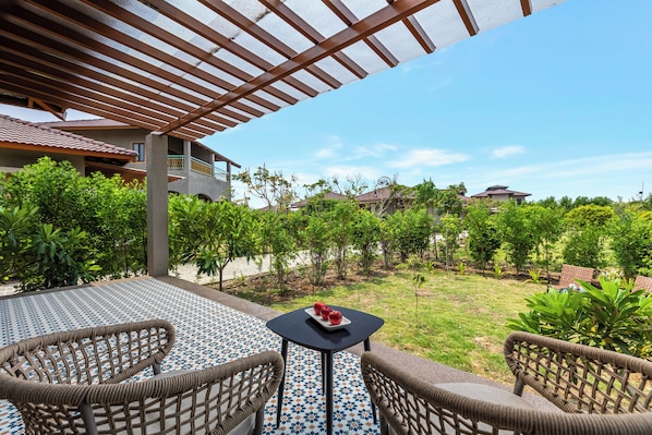 Uma área externa com assentos de vime e uma mesa pequena, com vista para um jardim repleto de plantas e um edifício ao fundo.