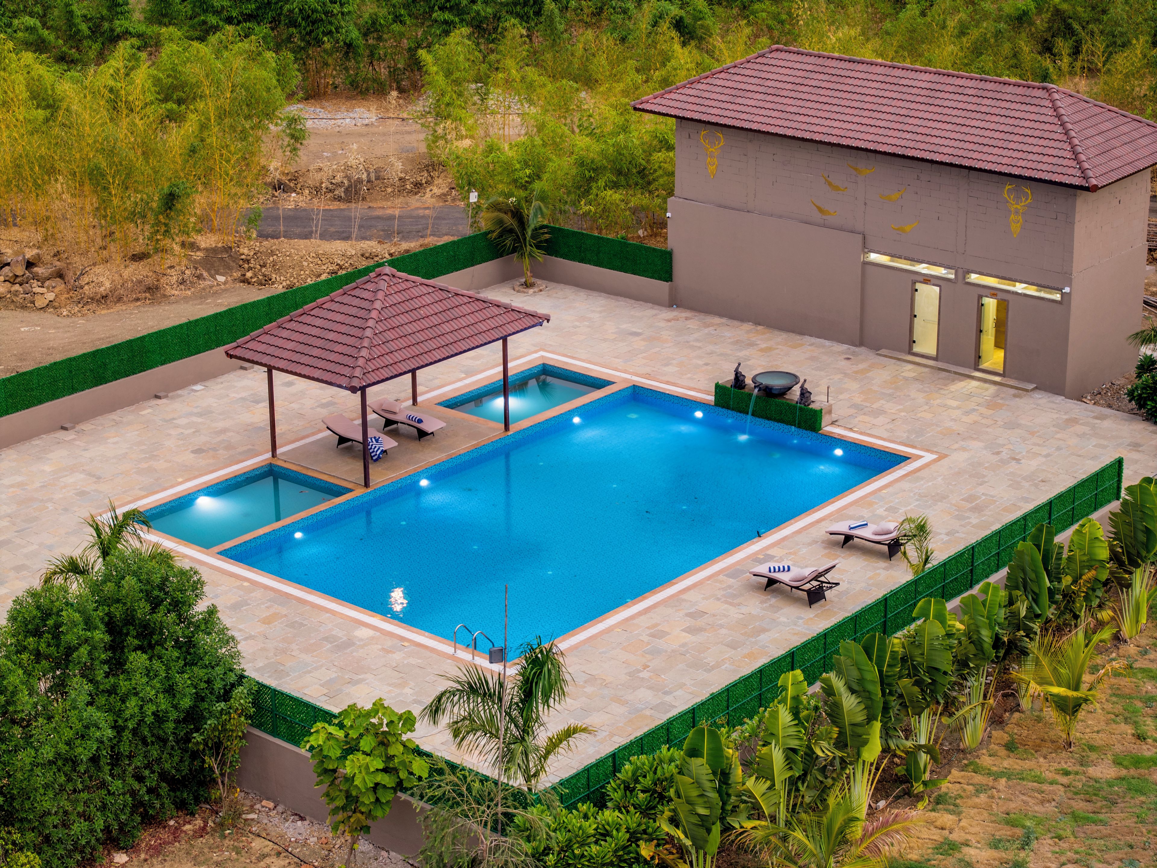 Piscina rectangular con zona cubierta y sillones de descanso, rodeada de follaje y un edificio pequeño con techo de tejas.