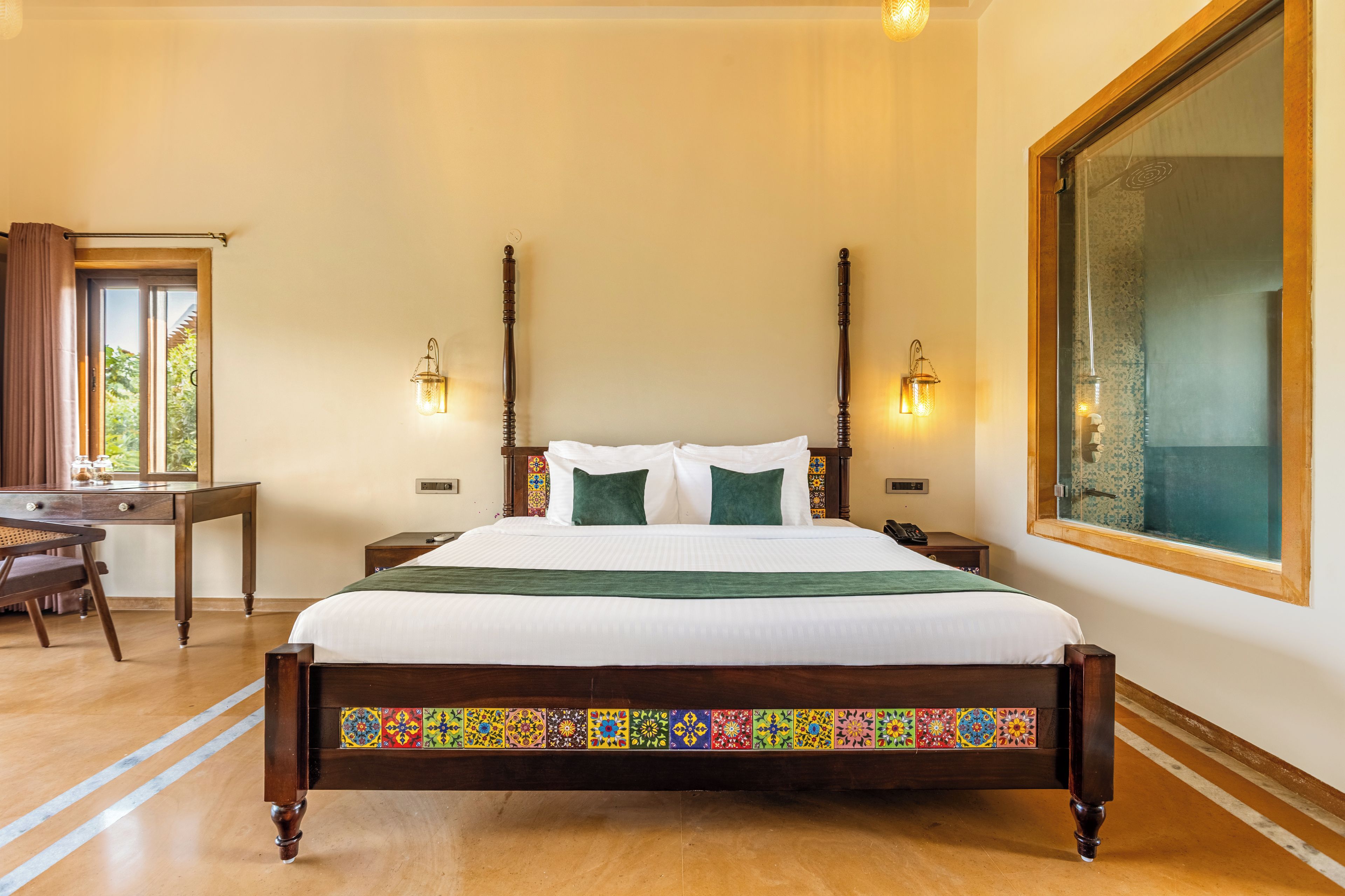 Una cama bien hecha con estructura de madera y detalles en cerámica de colores, un escritorio con silla y dos lámparas fijadas a la pared.