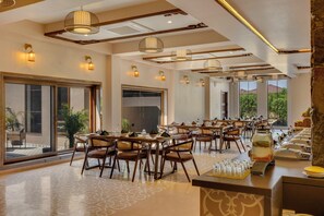A spacious dining area with multiple tables and chairs, large windows offering a view of the outdoors, and a buffet line with various food items.