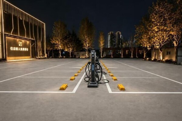 A parking area with a charging station, trees, and a building with illuminated signage. - QING LAI IMAGE ART HOTEL Heda New District(YOUXI HOTEL) (Baoding)