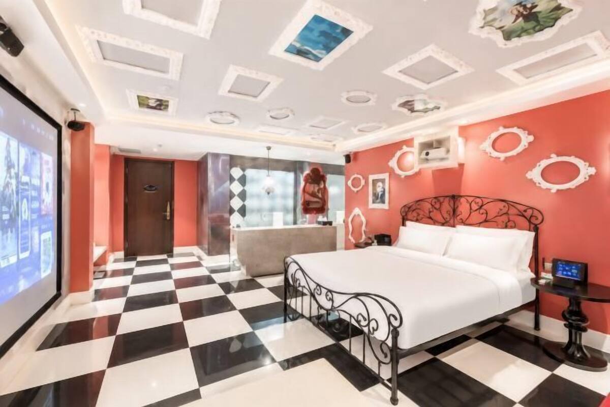 Una habitación de hotel con piso a cuadros, una cama con estructura de metal negro, una pared de acento roja y un techo con luces empotradas.