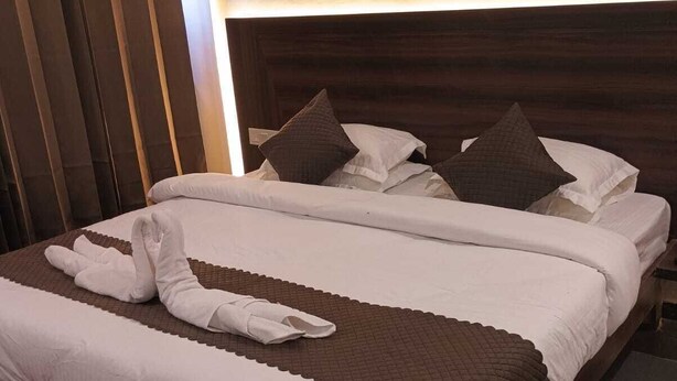 A neatly made bed with white linens and brown pillows, a wooden headboard, and a dark curtain. - Hotel Amber (Jaipur)