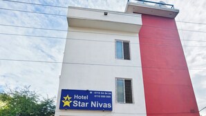 Un edificio de dos pisos con fachada roja y blanca, un cartel que dice 'Hotel Star Nivas' con números de contacto, y un área de entrada pavimentada con plantas en macetas.