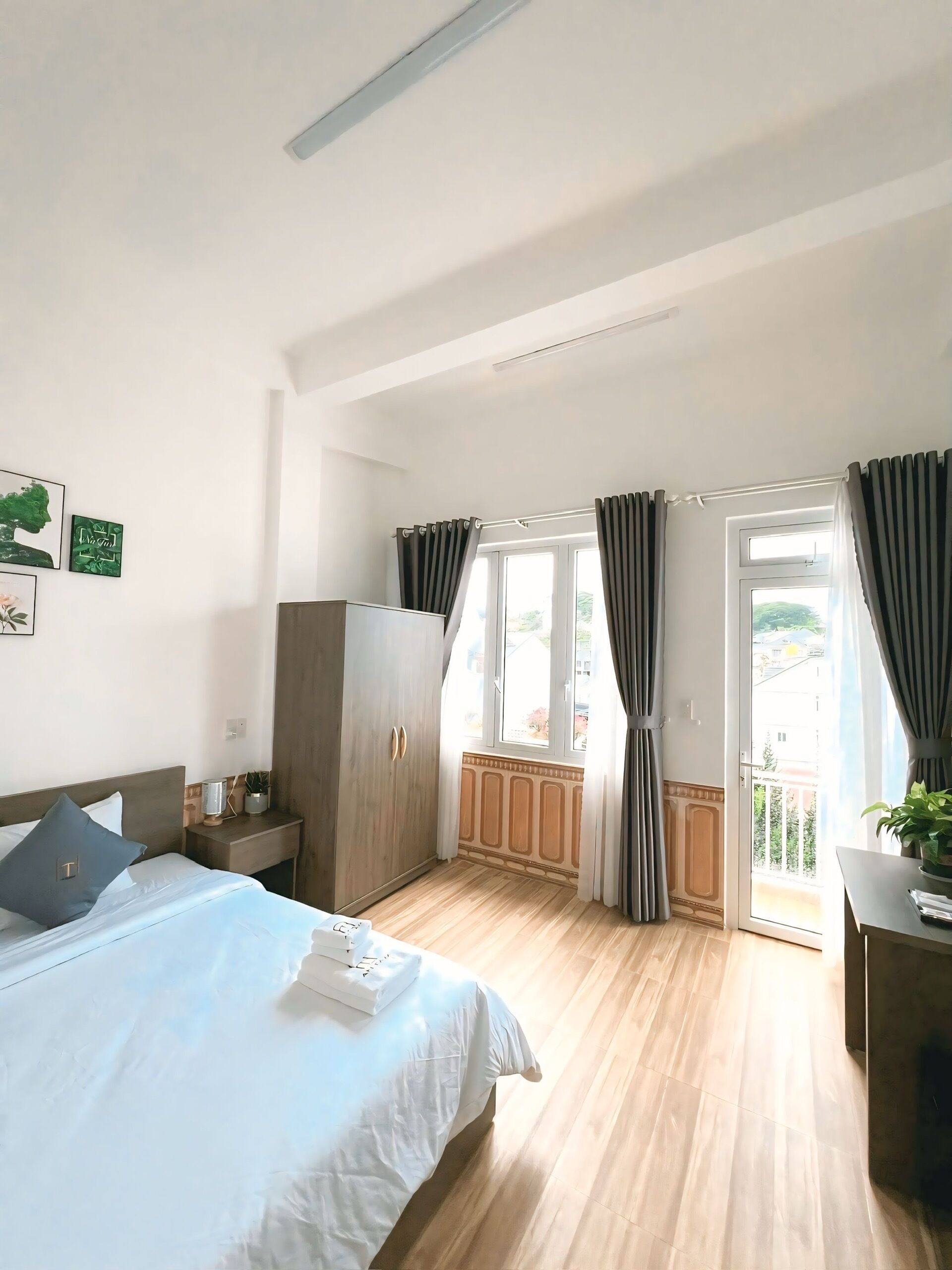 Ein Schlafzimmer mit Bett, Kleiderschrank, Nachttisch mit Lampe, Fenster mit Vorhängen und Blick ins Freie.