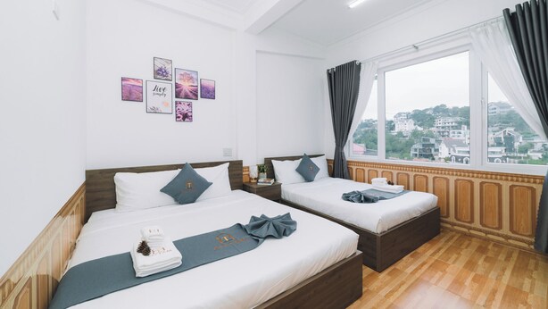Habitación de hotel con dos camas, un ventanal con cortinas, vistas a la ciudad y una pared decorada con cuadros.