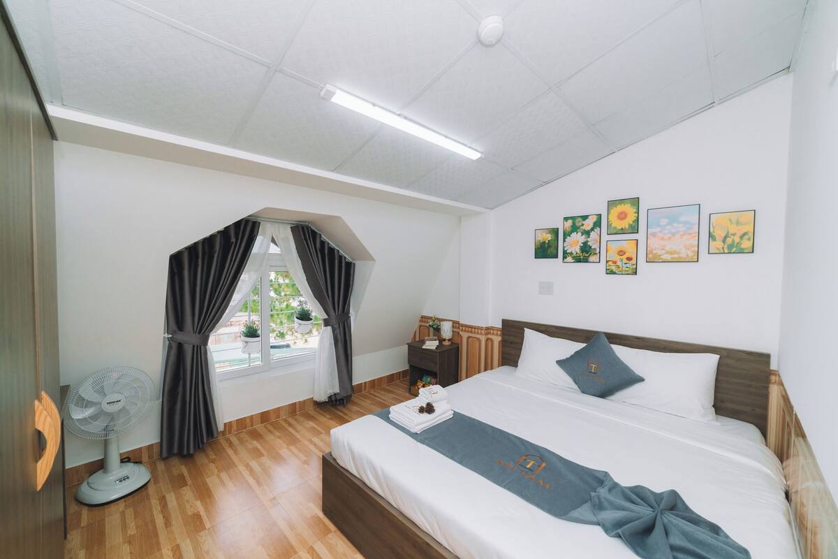 A hotel room with a bed, a bedside table, a nightstand, a window with curtains, and wall art.
