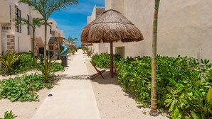 A pathway lined with palm trees, a thatched umbrella, and a building with a modern design.