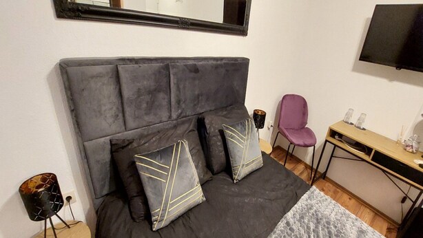 Ein Schlafzimmer mit einem dunkelgrauen Bett, zwei Kissen mit geometrischen Mustern, einem kleinen runden Tisch, einem violetten Sessel, einem Holztisch und einem an der Wand befestigten Flachbildfernseher.