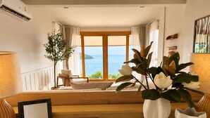Een kamer met uitzicht op zee, een bank, een stoel, een tafel met een lamp en twee vazen met bloemen.