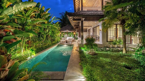 Uma piscina com espreguiçadeiras, uma casa com grandes janelas e vegetação exuberante.