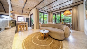 Uma sala de estar moderna com sofá, mesa de centro redonda, área de cozinha e janelas amplas com cortinas.