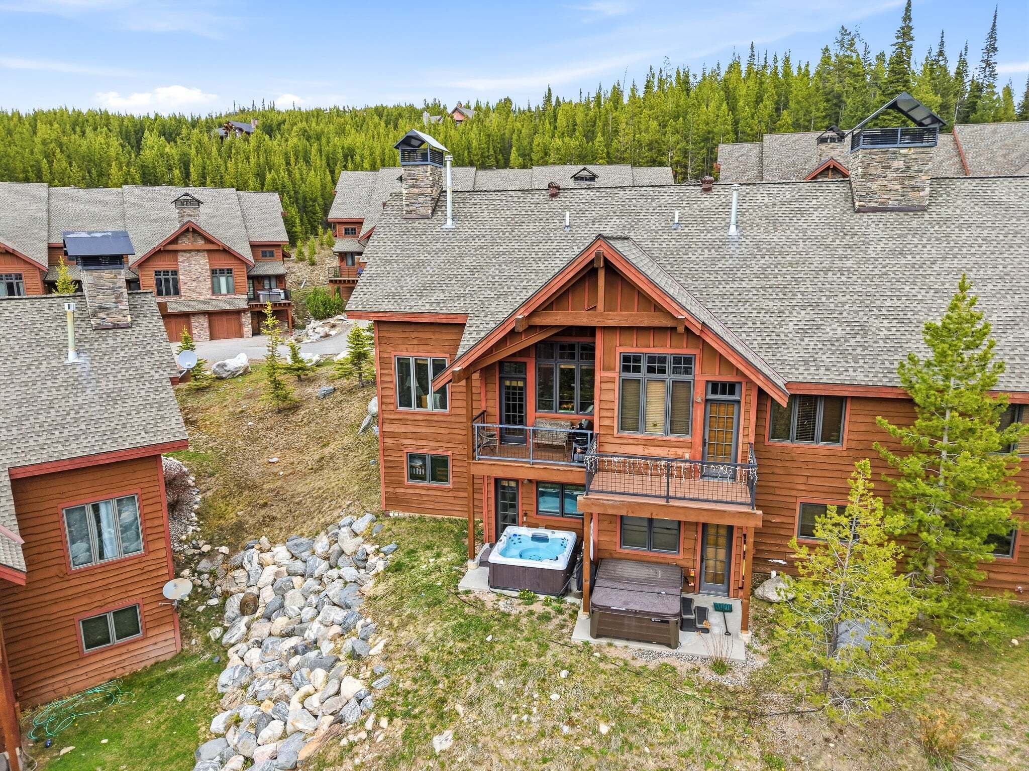 A multi-story lodge with a stone facade, a hot tub, and a view of a forested hillside.