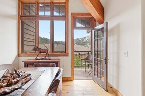 Ein Zimmer mit einem Holztisch, einer Statue, großen Fenstern mit Bergblick und einer Glastür, die nach draußen führt.