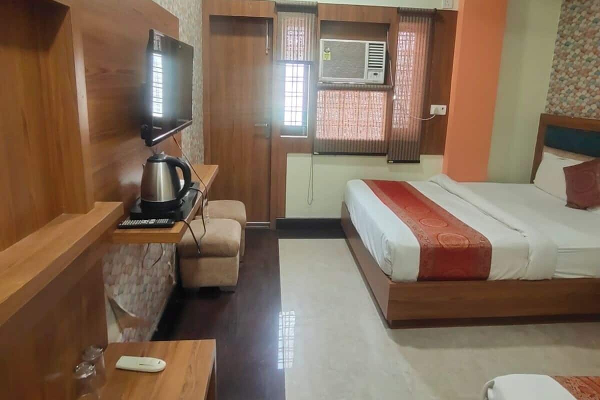 A hotel room with a bed, a TV, a kettle, a small table, and a window with curtains.