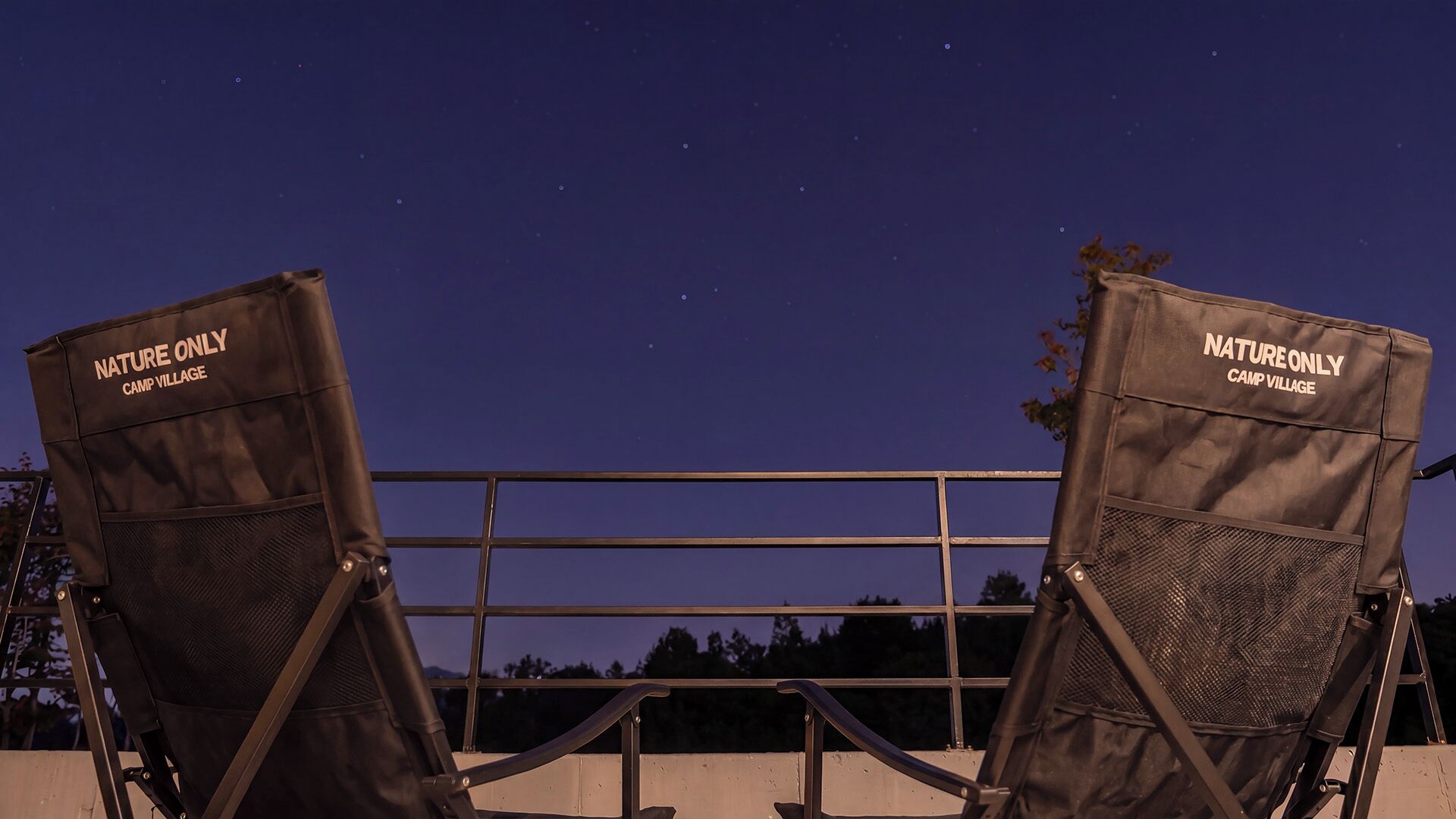 Two nature-themed lounge chairs with 'NATURE ONLY CAMP VILLAGE' branding, positioned on a balcony with a metal railing, overlooking a night sky.