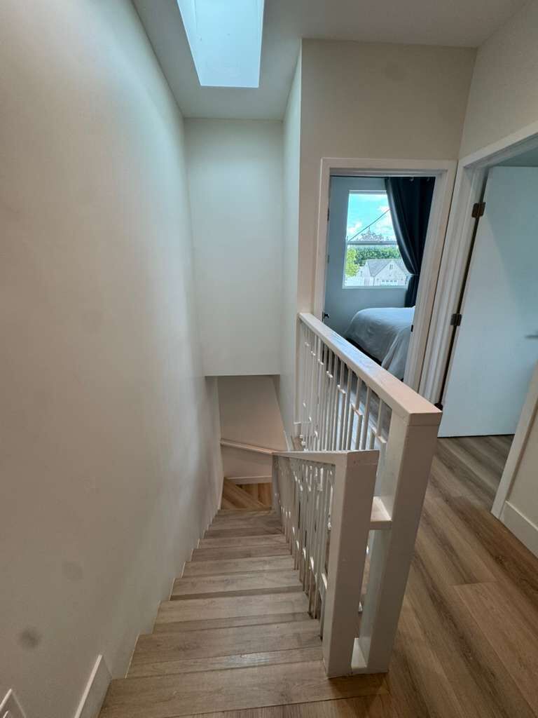 Unes escales estretes de fusta amb baranes blanques, que condueixen a una habitació amb finestra i porta.