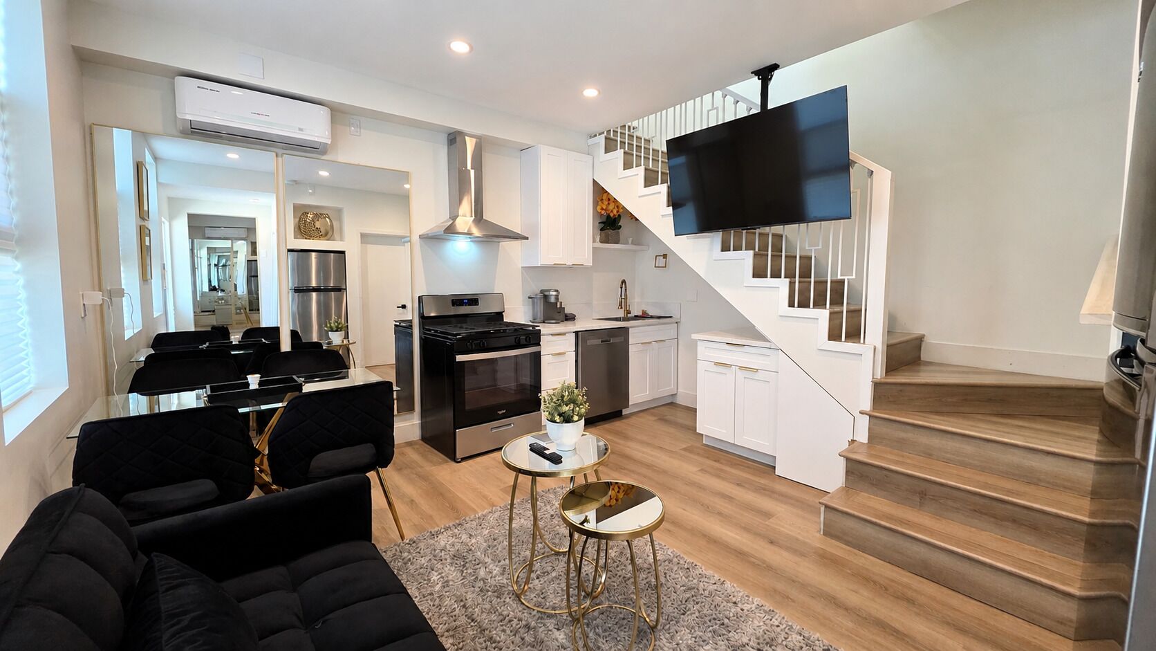 Una sala moderna con un sofá, una mesa de centro y una escalera que lleva a una cocina con armarios blancos y un televisor negro.