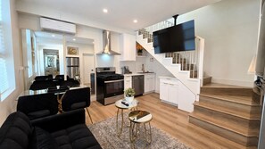 A modern living room with a sofa, a coffee table, and a staircase leading to a kitchen with white cabinets and a black TV.