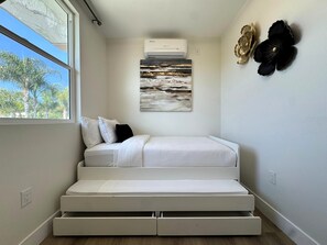 A compact room with a bed, a window, an air conditioner, and a wall-mounted artwork.