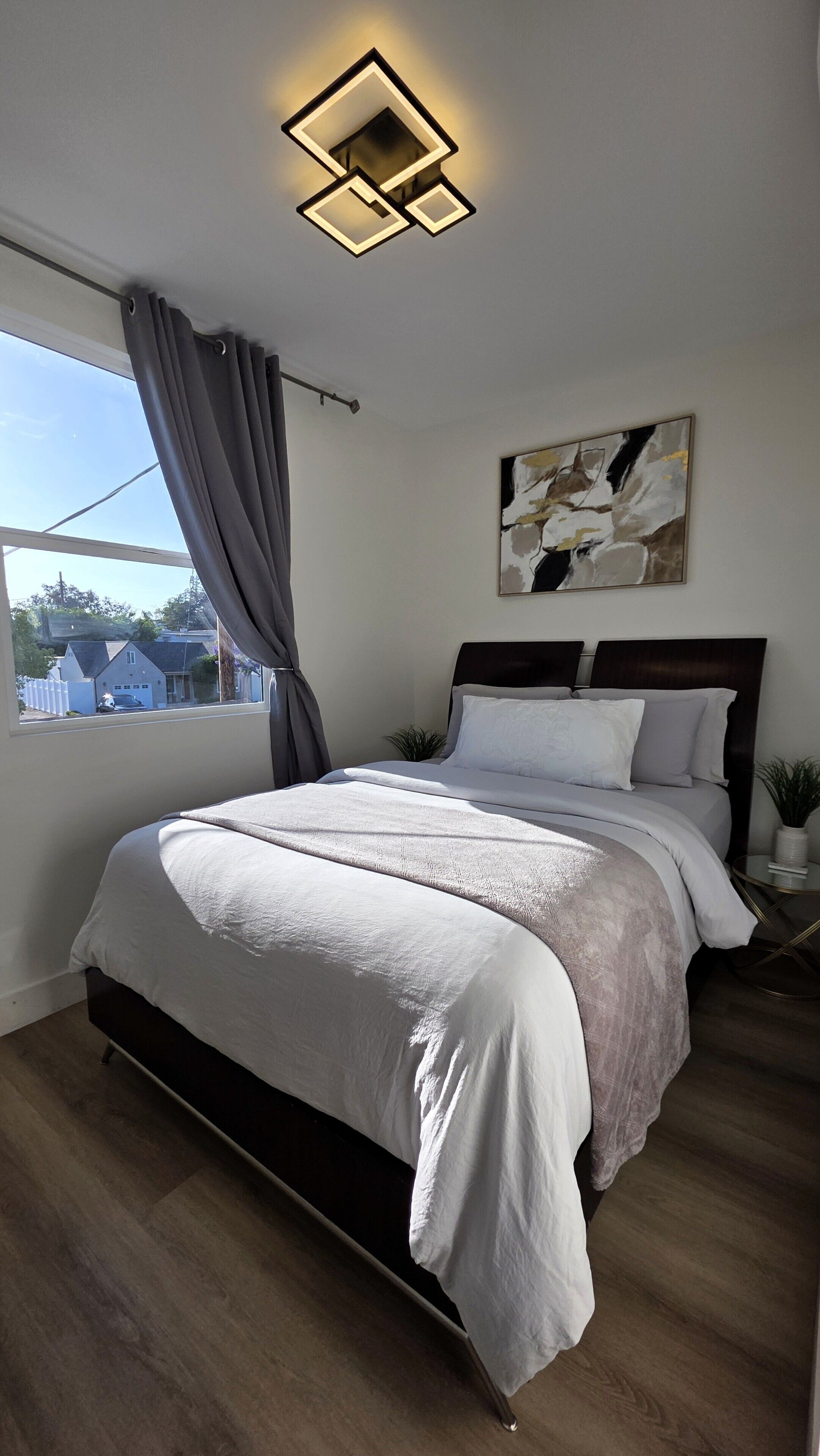 A modern bedroom with a bed, a painting, a window with curtains, and a ceiling light fixture.