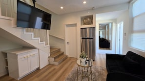A modern living room with a staircase, a flat-screen TV, a refrigerator, and a black sofa.