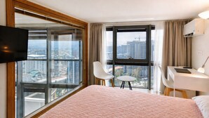 Ein Hotelzimmer mit einem großen Bett, einem Schreibtisch, zwei Stühlen, einem Fernseher und großen Fenstern mit Blick auf die Stadt.