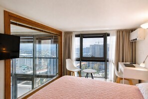 Ein Hotelzimmer mit einem großen Bett, einem Schreibtisch, zwei Stühlen, einem Fernseher und großen Fenstern mit Blick auf die Stadt.