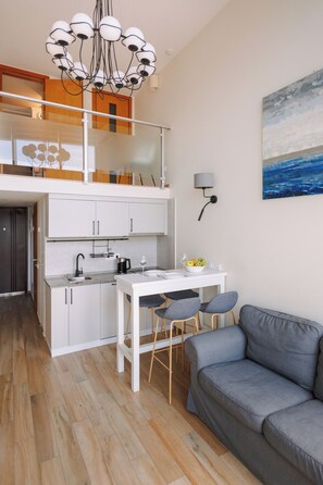 Eine moderne Küche mit weißen Schränken, einem Esstisch mit zwei Stühlen, einem grauen Sofa und einer Pendelleuchte.