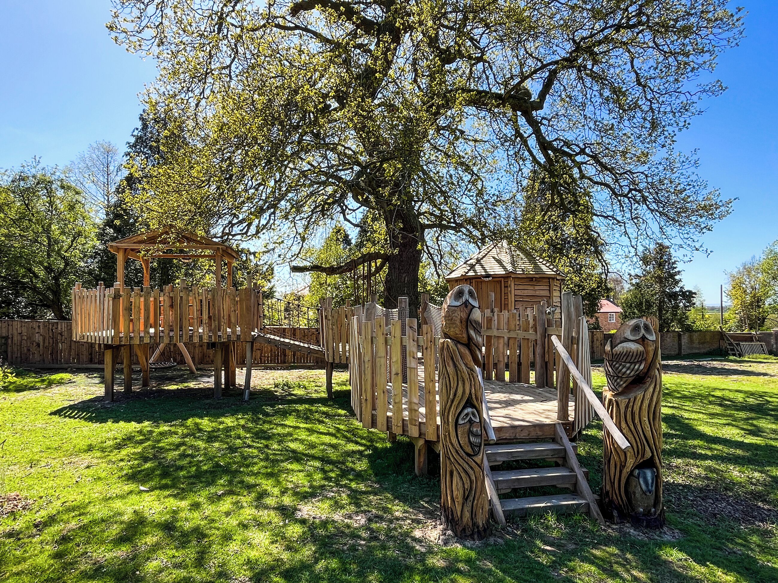 A playground with wooden structures, including a treehouse and a slide, surrounded by grass and trees.
