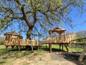 Eine hölzerne Spielkonstruktion mit einer Seilbrücke und einem kleinen Holzhaus, eingebettet in eine grüne Wiese mit einem großen Baum.