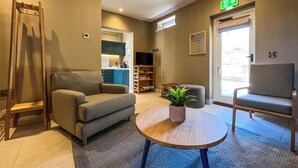 Een hotellobby met een bank, een kleine tafel met een plant, een stoel, een trap en een kitchenette op de achtergrond.