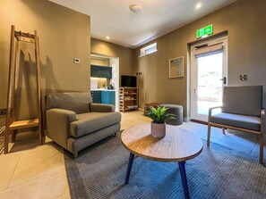 Eine Hotelhalle mit einer Couch, einem kleinen Tisch mit Pflanze, einem Sessel, einer Treppe und einer Küchenzeile im Hintergrund.