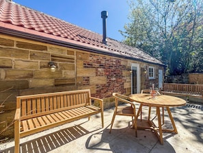 A stone building with a red-tiled roof, a wooden bench, and a round wooden table with chairs.