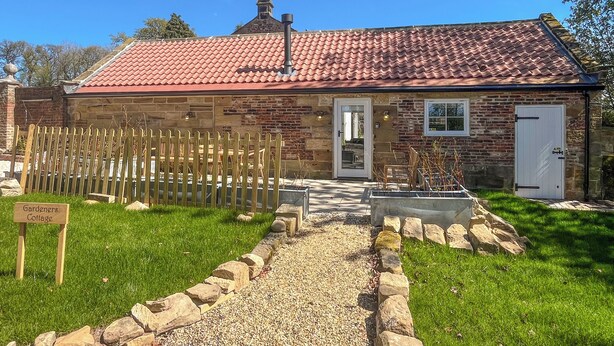 A stone cottage with a red-tiled roof, a wooden fence, and a gravel path leading to the entrance.