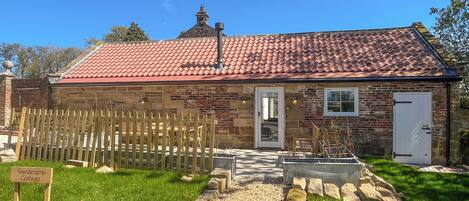 A stone cottage with a red-tiled roof, a wooden fence, and a gravel path leading to the entrance.