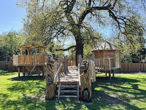 A wooden play structure with a rope bridge, a wooden house, and a tree in a park.