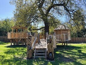 Eine Spielanlage aus Holz mit einer Hängebrücke, einem Holzhaus und einem Baum in einem Park.
