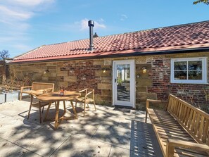 A stone building with a tiled roof, a wooden table with chairs, and a bench.