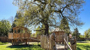 Un terrain de jeux doté de structures en bois, notamment une cabane dans les arbres et un toboggan, entouré d’herbe et d’arbres.