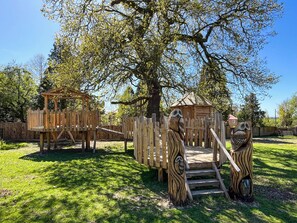 Ein Spielplatz mit Holzkonstruktionen, darunter ein Baumhaus und eine Rutsche, umgeben von Gras und Bäumen.