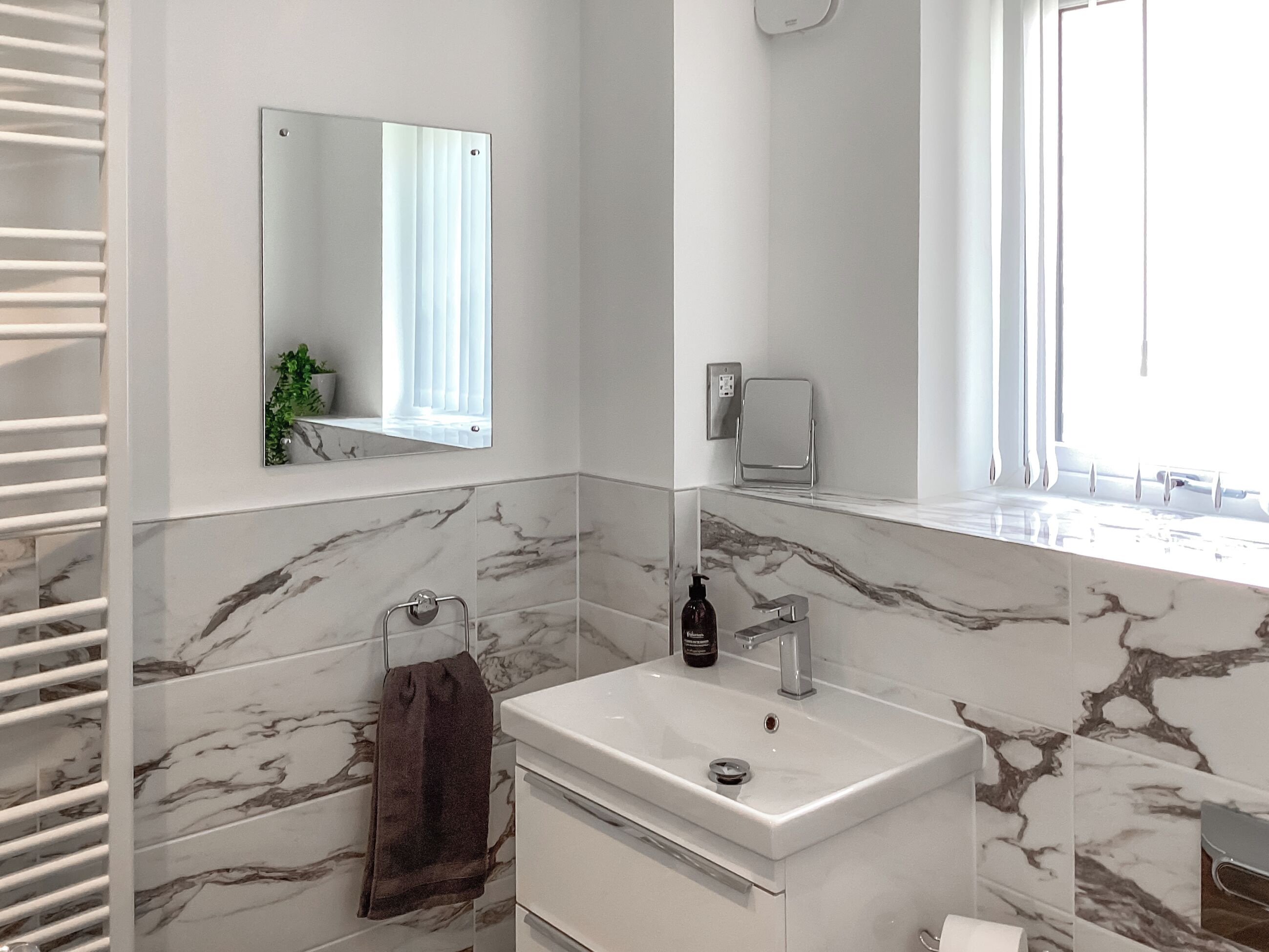 A bathroom with a marble-tiled wall, a white sink, a towel rack, a mirror, and a window with blinds.