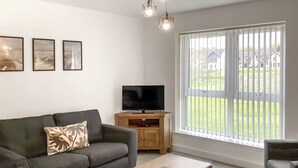 Ein Wohnzimmer mit einer Couch, einem Couchtisch, einem Fernseher auf einer Holzkommode und einem großen Fenster mit Jalousien.