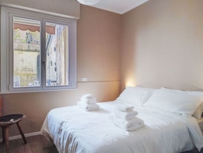 Quarto de hotel com uma cama, uma cadeira e uma janela com vista para um edifício e árvores.