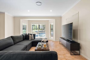 Ein Wohnzimmer mit einem Ecksofa, einem Couchtisch, einem Flachbildfernseher und einem großen Fenster mit Blick auf Bäume.