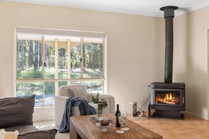 Ein gemütliches Wohnzimmer mit großem Fenster, einer Couch, einem Holztisch mit Vase, einer Weinflasche und Gläsern, sowie einem Kaminofen.