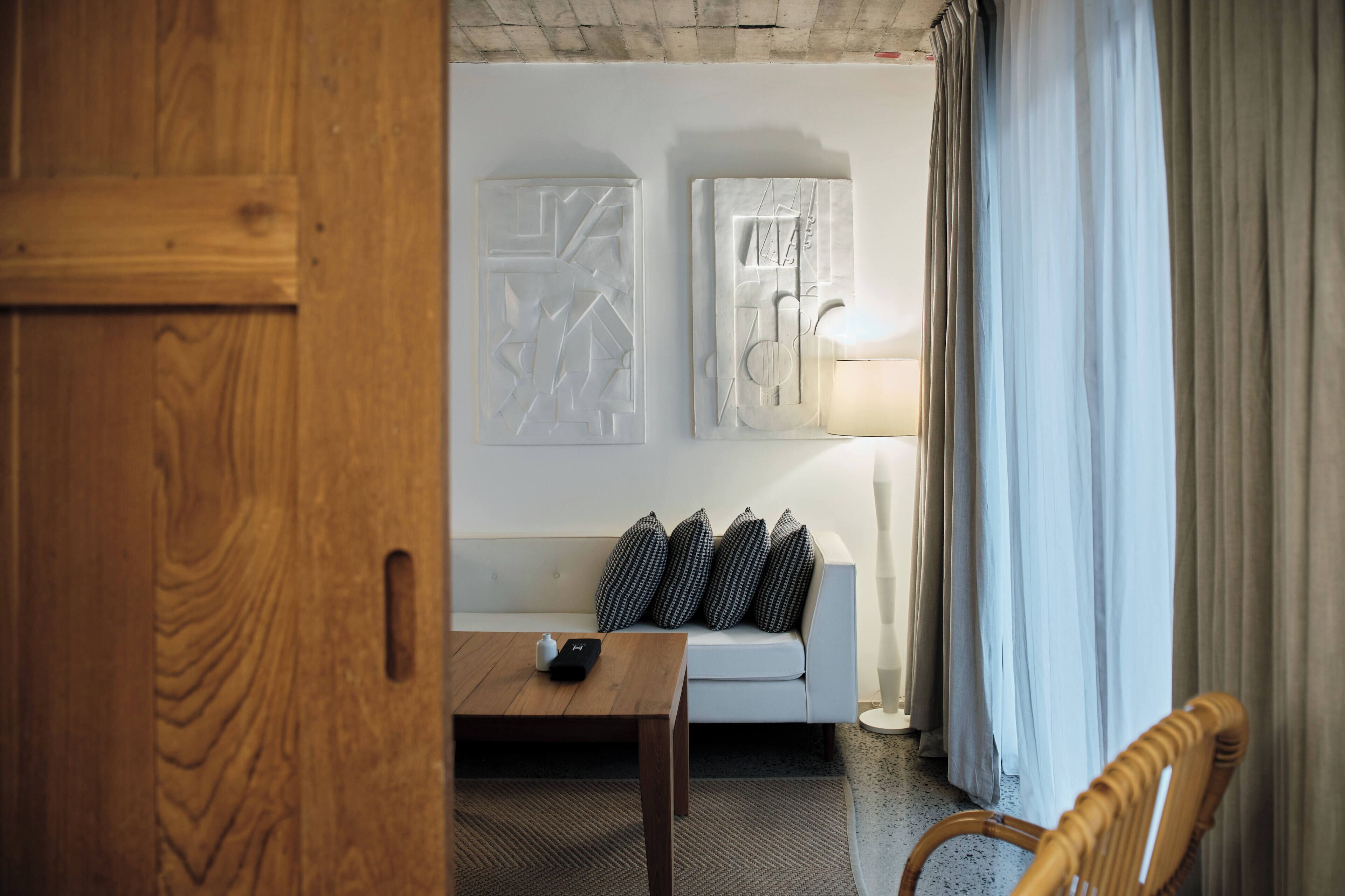 A room with a wooden door, a white sofa with patterned cushions, a small table with a remote control, and a lamp.