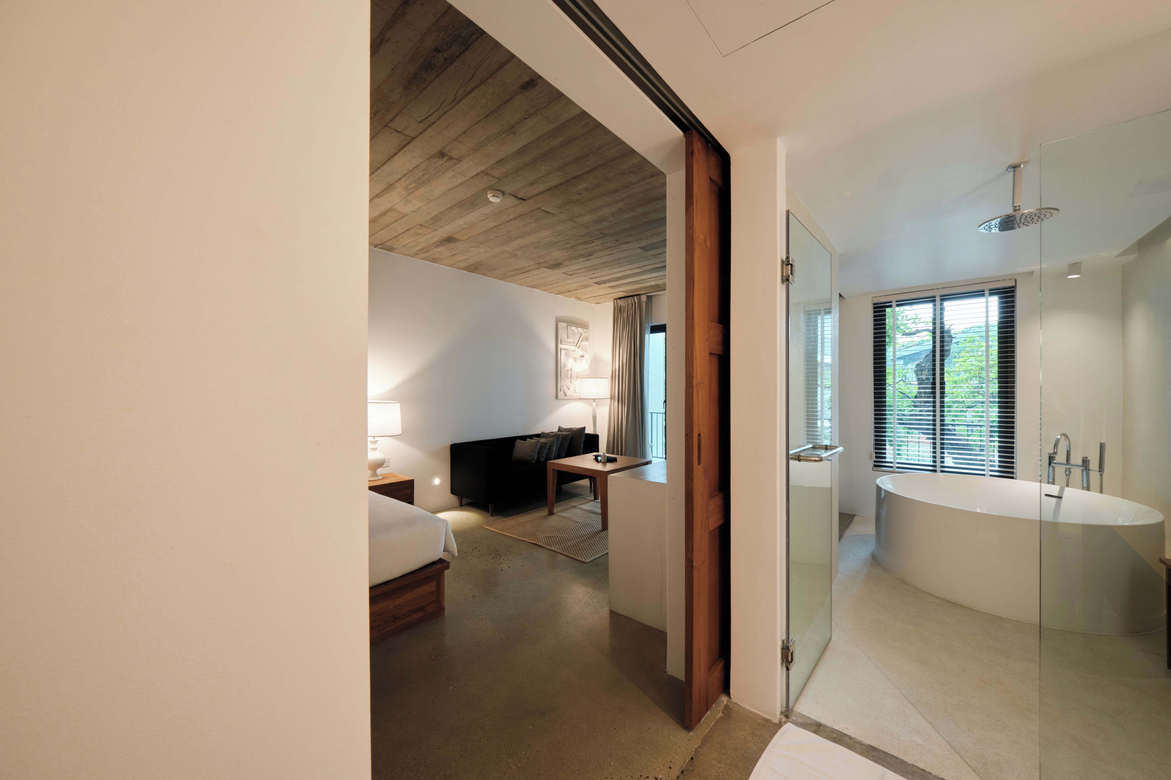 A modern hotel room with a wooden ceiling, a glass-enclosed bathroom, a large bathtub, and a seating area with a sofa and table.