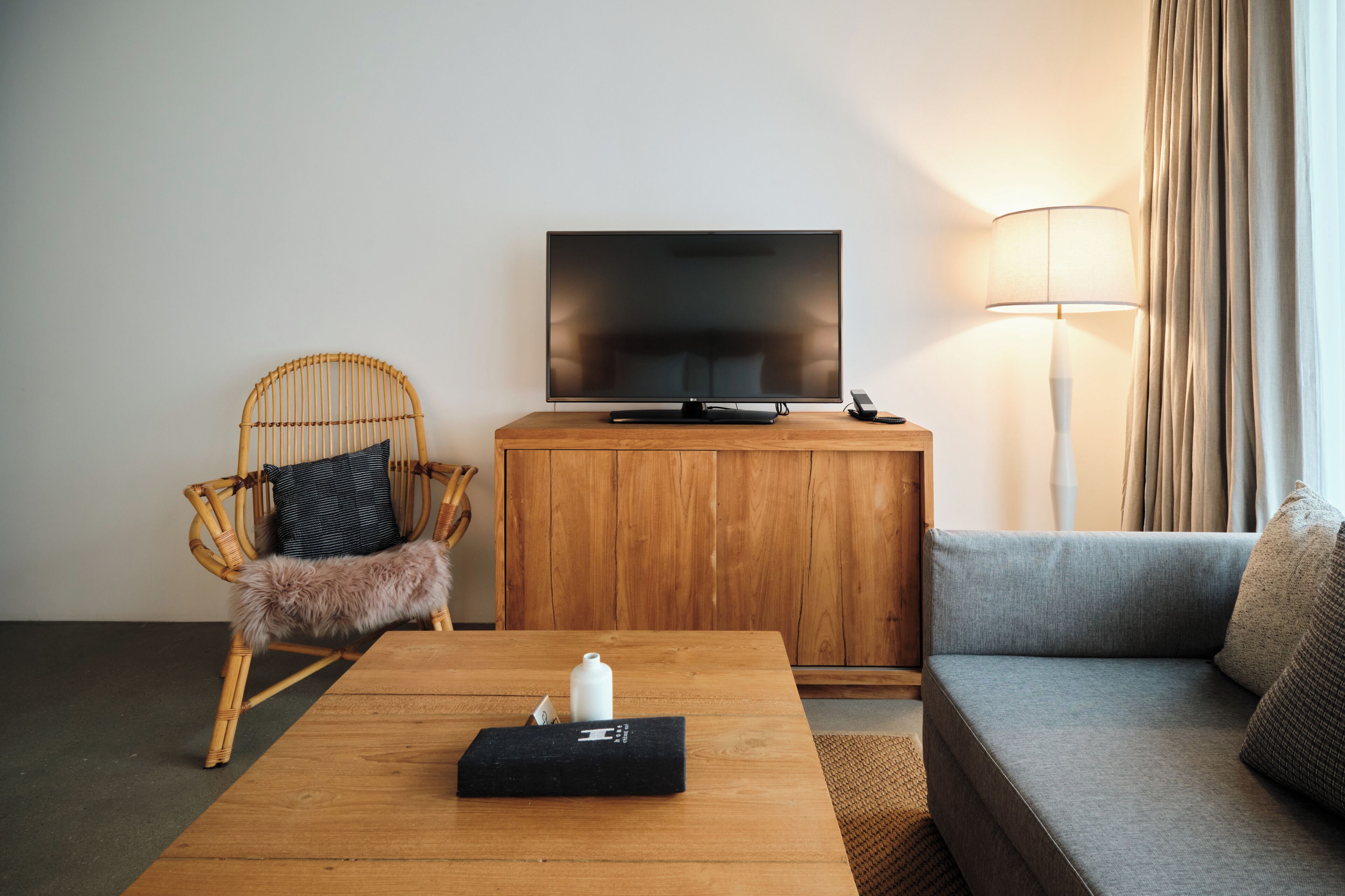 Quarto de hotel com televisão de tela plana, um armário de madeira, uma poltrona de vime com manta de pele, um sofá cinza e um abajur.