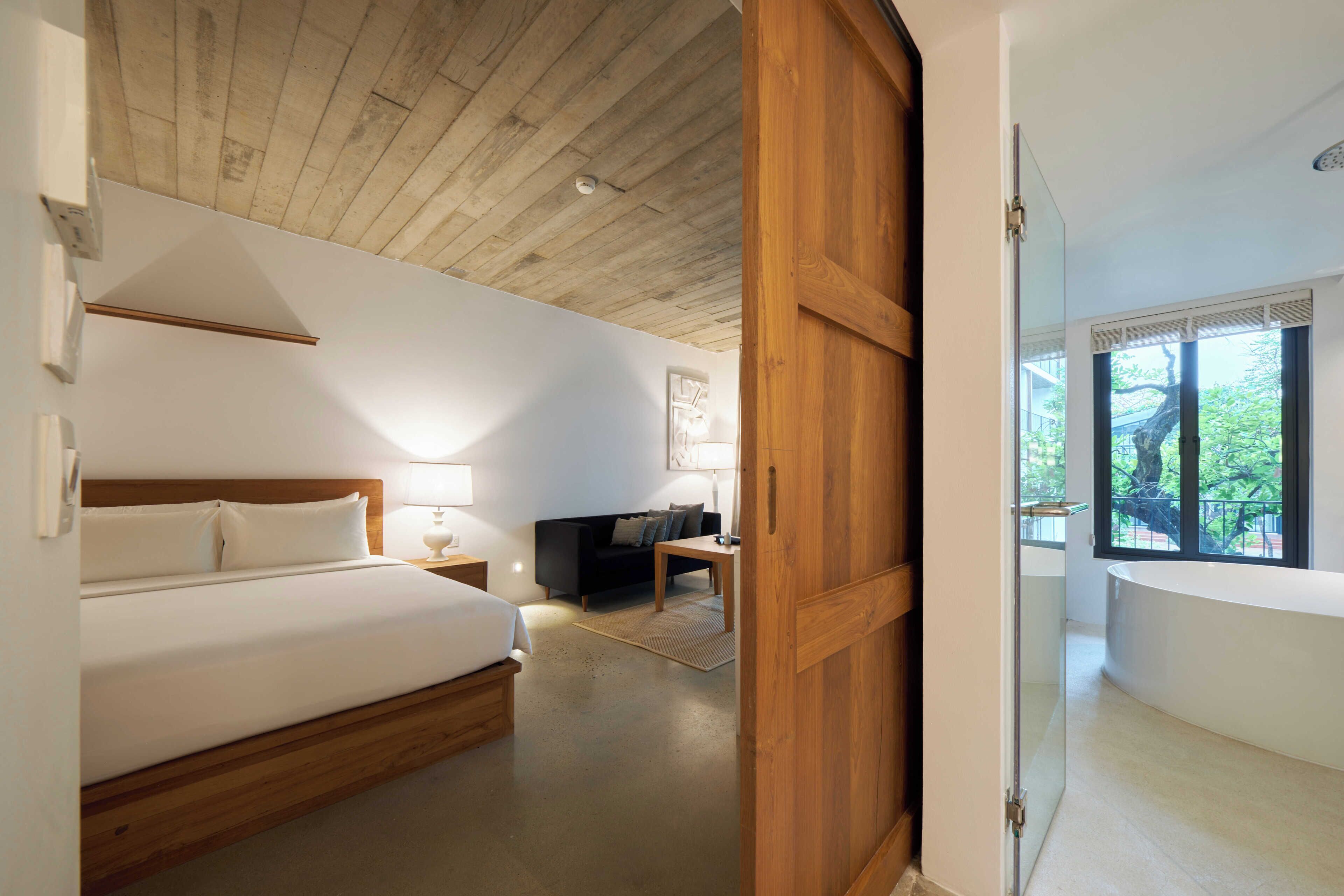 A bedroom with a wooden ceiling, a bed, a nightstand, a sofa, and a view of a bathroom with a bathtub.